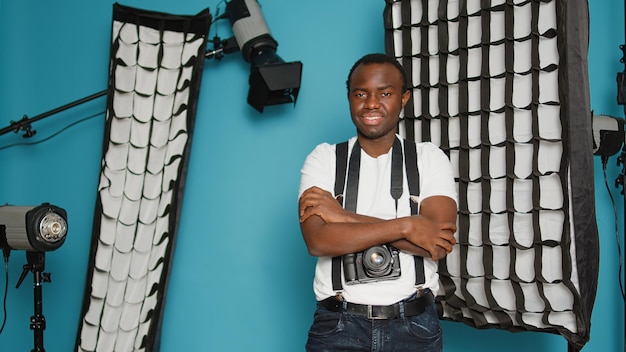 Photographe masculin posant les bras croisés dans les coulisses du studio, disposant d'un équipement de photographie professionnel. Utilisation des outils d'éclairage, de réflecteur, de trépied et de boîte à lumière des coulisses.