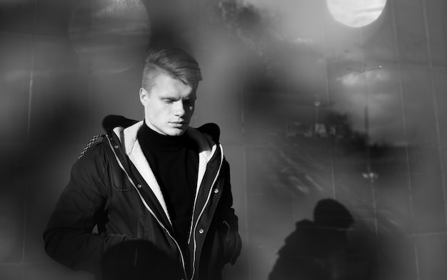 Photographie artistique en noir et blanc. Jeune homme blond en tenue noire debout dans la rue.
