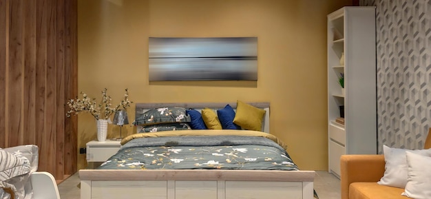 Photographie d'intérieur grande chambre avec grand lit intérieur de chambre à coucher avec drap de lit ton jaune et image sur le mur tête de lit en bois de style maison douce