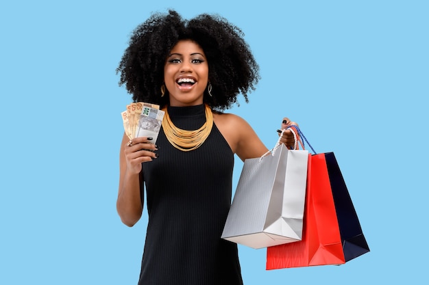 Photographie d'une jeune femme afro souriante tenant des sacs de courses dans une main et de l'autre tenant des billets d'argent brésiliens isolés sur un fond cyan pastel