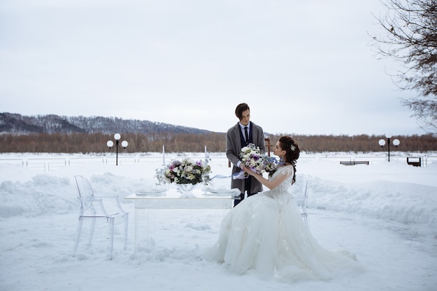 Photographie de mariage mise en scène en hiver. Jeunes mariés avec un beau bouquet à côté d'une table et de chaises en verre