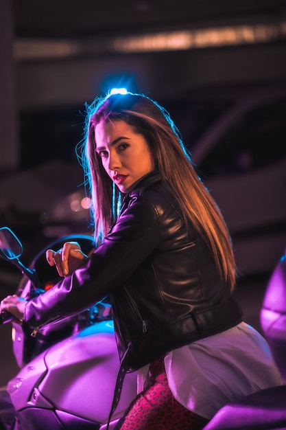 Photographie avec des néons bleus et roses sur une moto par derrière. Portrait d'une jeune mannequin blonde caucasienne portant une veste en cuir noir