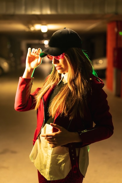 Photographie avec néons rouges et verts dans un parking. Jeune femme de race blanche jolie blonde dans un costume rouge, des lunettes de soleil et une casquette noire