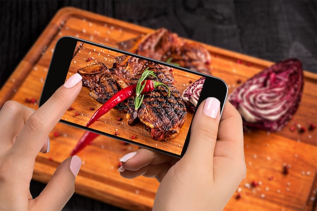 Photographier le concept de nourriture - le touriste prend une photo d'un plat de steak de boeuf prêt à manger sur une planche de bois à découper sur un smartphone. Service original de nourriture au restaurant