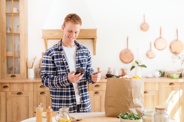 Épicerie en ligne Un homme utilisant une carte de crédit et un téléphone paie pour des produits alimentaires debout dans la cuisine à la maison