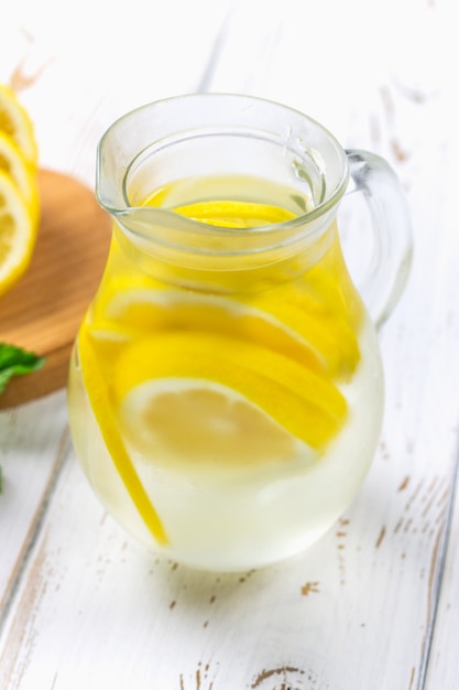 Un pichet avec une limonade froide sur un fond en bois blanc entouré de citrons.