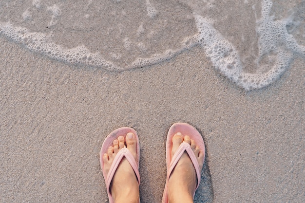 Pieds de femme portent des pantoufles debout sur la plage tropicale de sable