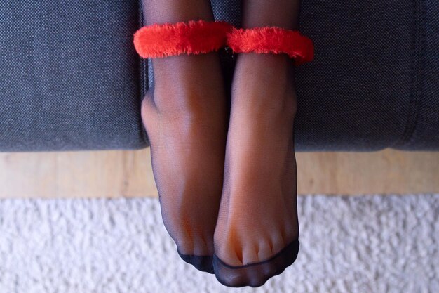 Photo des pieds de femmes avec des menottes rouges en peluche dans des collants noirs sur un canapé gris