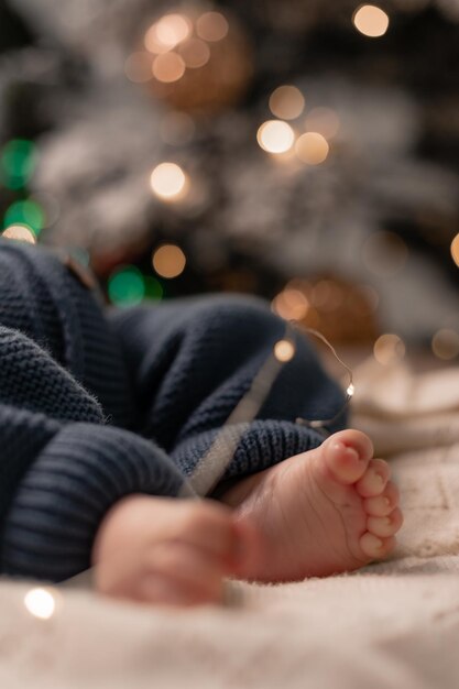 Pieds nus d'un bébé dans une combinaison tricotée bleue allongée sur une couverture légère sur le sol sous le gros plan de l'arbre de Noël. vacances d'hiver, moments drôles, noël enfant. Photo de haute qualité