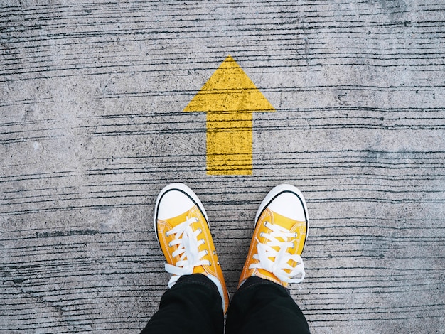 Pieds de selfie portant des baskets jaunes devant la flèche sur une route en béton.