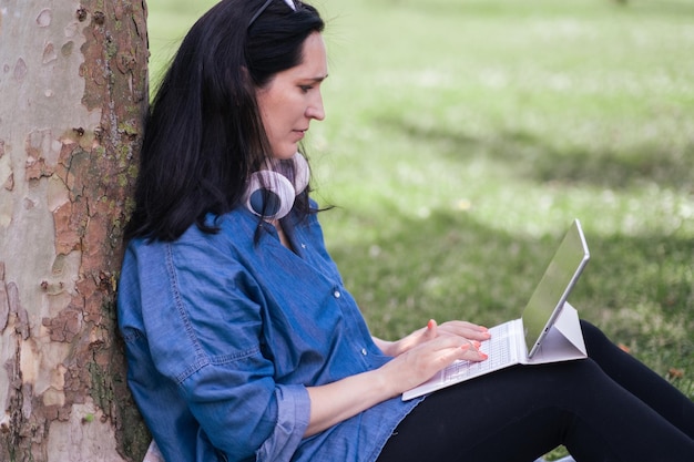 pigiste en chemise en jean communiquant avec l'employeur dans le chat en ligne via une tablette assise près d'un arbre