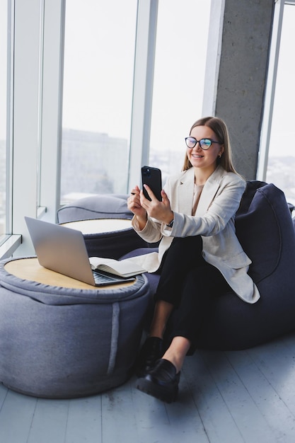 Une pigiste concentrée assise dans un espace de travail moderne avec un netbook ouvert et vérifiant les notifications sur un téléphone portable dans des vêtements décontractés
