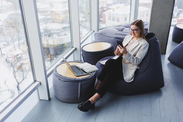 Une pigiste concentrée assise dans un espace de travail moderne avec un netbook ouvert et vérifiant les notifications sur un téléphone portable dans des vêtements décontractés