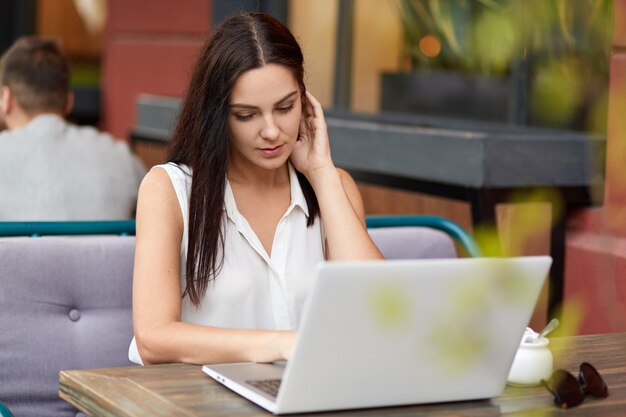 Une pigiste concentrée regarde un didacticiel en ligne, utilise un ordinateur portable pour travailler, s'assoit à table dans une cafétéria extérieure, attend sa commande. Femme fait du shopping sur le site Web avec des remises