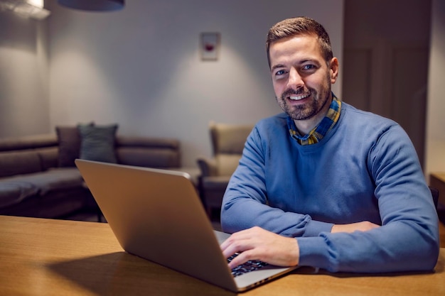 Un pigiste heureux travaillant en ligne sur l'ordinateur portable de la maison et souriant à la caméra