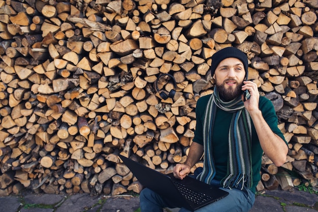 Pigiste d'homme d'affaires barbu dans des écouteurs travaillant sur un ordinateur portable et parlant au téléphone