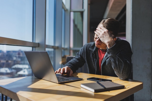 Un pigiste à lunettes regarde dans un ordinateur portable et il est en colère alors qu'il est assis à une table avec un ordinateur portable et un bloc-notes dans un café pendant la journée Journée malchanceuse Travail à distance depuis le bureau