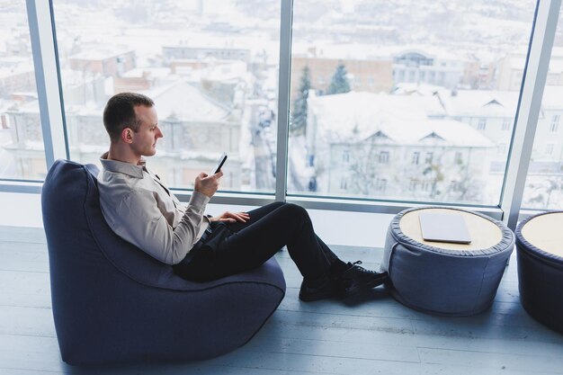 Pigiste masculin prospère assis dans un espace de travail moderne et vérifiant les notifications sur son téléphone portable dans des vêtements décontractés Travail à distance