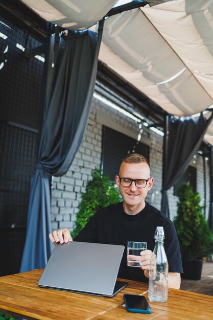 Un pigiste masculin prospère dans un t-shirt noir et des lunettes est assis sur la terrasse d'été d'un café et travaille sur un ordinateur portable Le directeur est assis dans un café pendant le déjeuner et travaille en ligne
