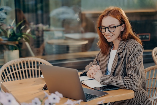 Une pigiste rousse regarde le webinaire de formation prend des notes dans le bloc-notes est assise devant un ordinateur portable ouvert habillé formellement pose dans un café en plein air navigue sur Internet développe un nouveau projet