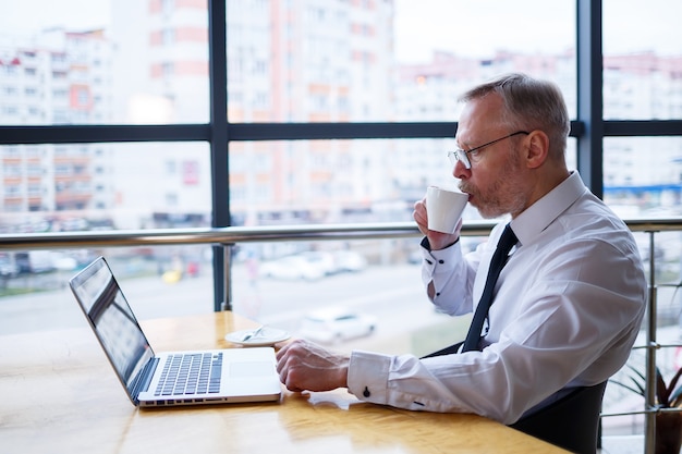 Un pigiste travaille dans un café sur un nouveau projet d'entreprise. Assis à une grande fenêtre à la table. Regarde un écran d'ordinateur portable avec une tasse de café