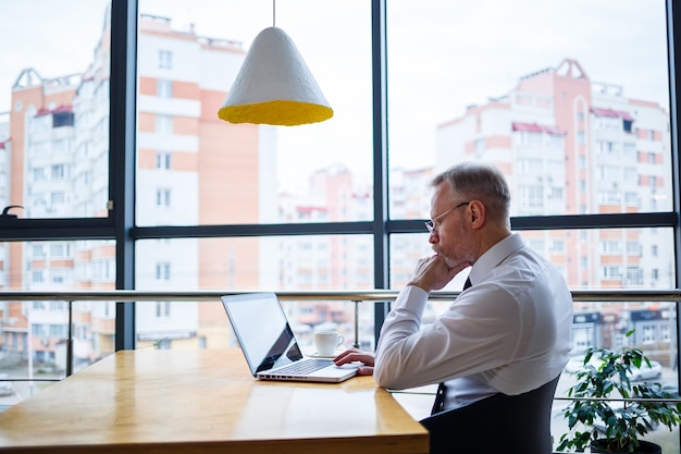 Un pigiste travaille dans un café sur un nouveau projet d'entreprise. Assis à une grande fenêtre à la table. Regarde un écran d'ordinateur portable avec une tasse de café