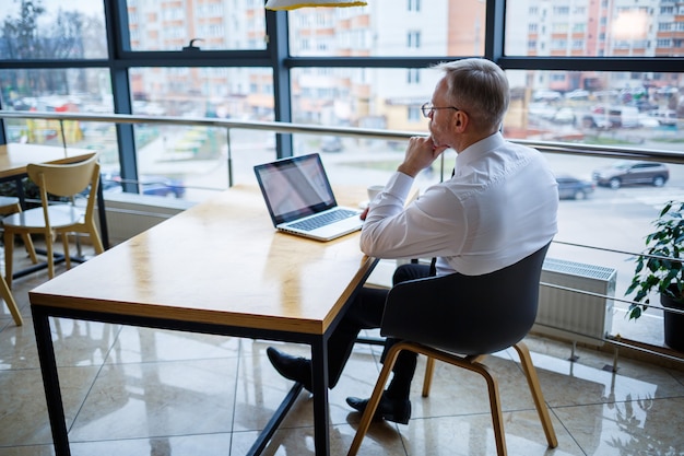 Un pigiste travaille dans un café sur un nouveau projet d'entreprise. Assis à une grande fenêtre à la table. Regarde un écran d'ordinateur portable avec une tasse de café