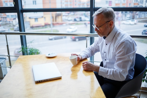 Un pigiste travaille dans un café sur un nouveau projet d'entreprise. Assis à une grande fenêtre à la table. Regarde un écran d'ordinateur portable avec une tasse de café