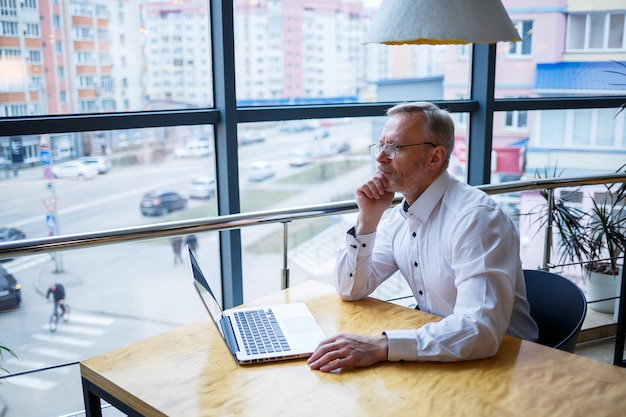 Un pigiste travaille dans un café sur un nouveau projet d'entreprise. Assis à une grande fenêtre à la table. Regarde un écran d'ordinateur portable avec une tasse de café