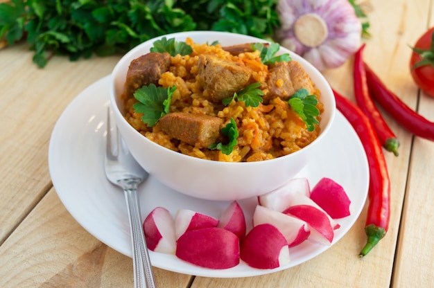 Photo pilaf asiatique traditionnel avec des herbes fraîches, de l'ail, des carottes et de la viande dans un bol blanc sur un fond en bois