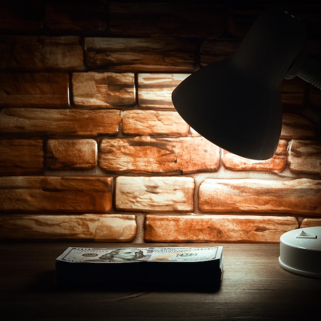 Une pile de billets de cent dollars sur une table en bois est éclairée par la lumière d'une lampe