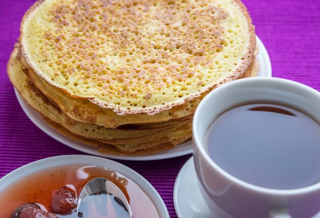 Pile de crêpes dorées avec confiture de fraises et tasse de thé. Fermer.