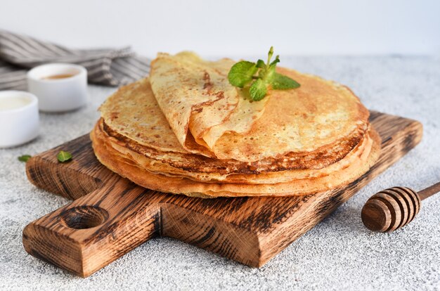 Une pile de crêpes fines avec de la crème sure et du miel sur une planche de bois.