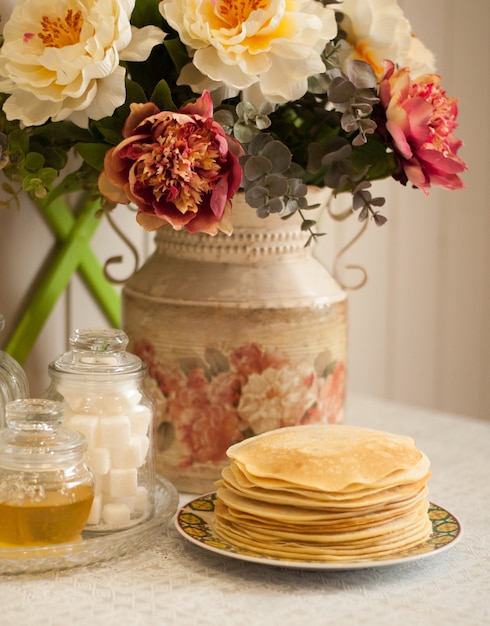 Une pile de crêpes sur fond de pivoines dans un vase et des plats