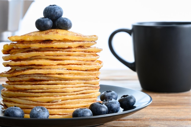 Pile de crêpes avec une tasse noire aux bleuets et une cafetière sur une table en bois