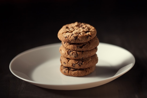 Pile de délicieux cookies aux pépites de chocolat sur une plaque blanche sur une vieille table en bois sombre. Biscuits maison.