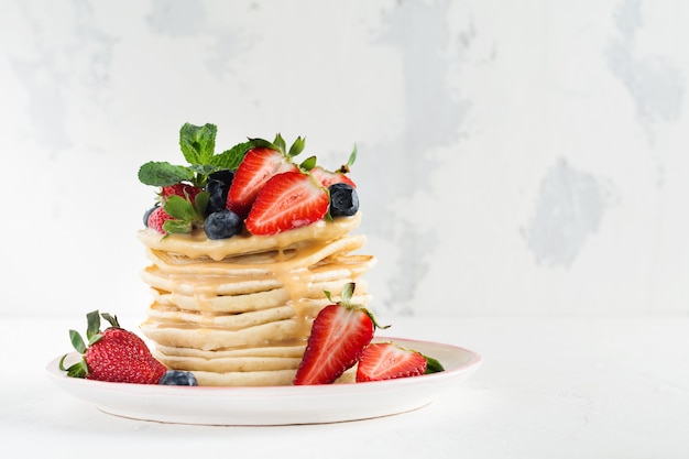 Pile de petites crêpes au miel, fraises fraîches et sauce sur fond en bois clair.