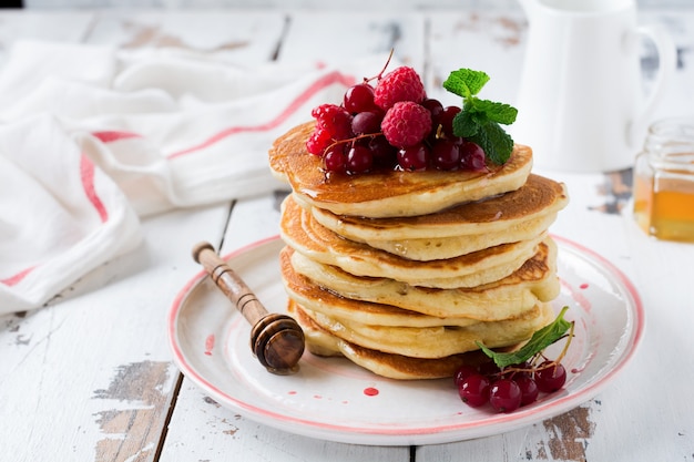 Pile de petites crêpes maison au miel, framboises fraîches et groseilles rouges sur une vieille surface en bois clair.