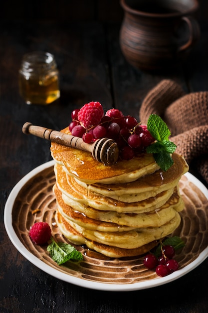 Pile de petites crêpes maison au miel, framboises fraîches et groseilles rouges sur une vieille surface en bois.