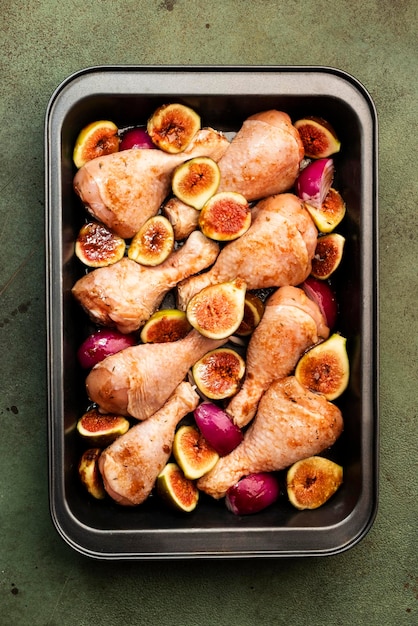 Pilons de cuisses de poulet crues avec figues douces et oignons rouges dans une marinade balsamique préparées pour la cuisson sur une plaque à pâtisserie vue de dessus sur fond vert rouillé