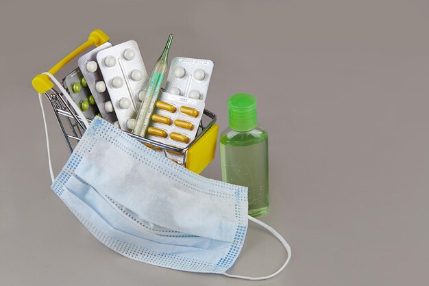 Photo des pilules, des médicaments, un thermomètre, un masque médical et un gel désinfectant dans un chariot de supermarché sur un fond gris photo de haute qualité