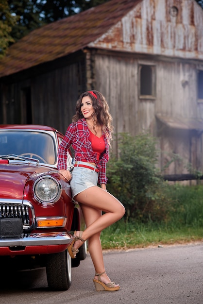 Pin-up femme posant près d'une voiture vintage rouge à la campagne.