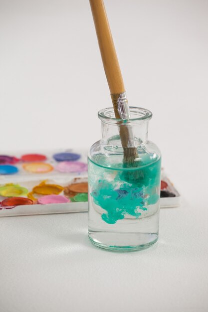 Pinceau avec de la peinture bleue trempée dans un pot rempli d'eau contre une surface blanche
