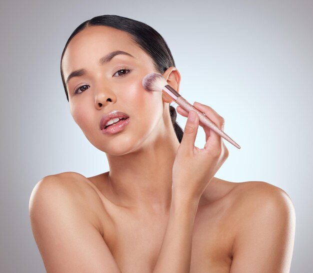 Les pinceaux de maquillage sont des baguettes magiques pour les femmes Portrait en studio d'une belle jeune femme se maquillant le visage avec un pinceau sur fond gris