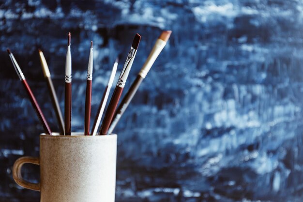 Pinceaux de peinture dans une tasse en céramique sur un fond abstrait bleu