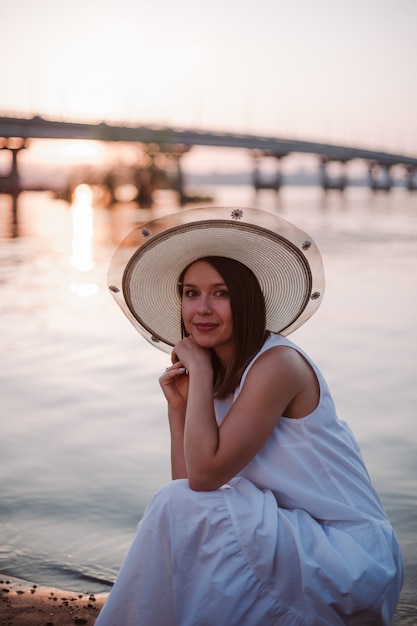 Pique-nique au coucher du soleil au pont sur la rivière femme détendue caucasienne dans une robe blanche et un chapeau