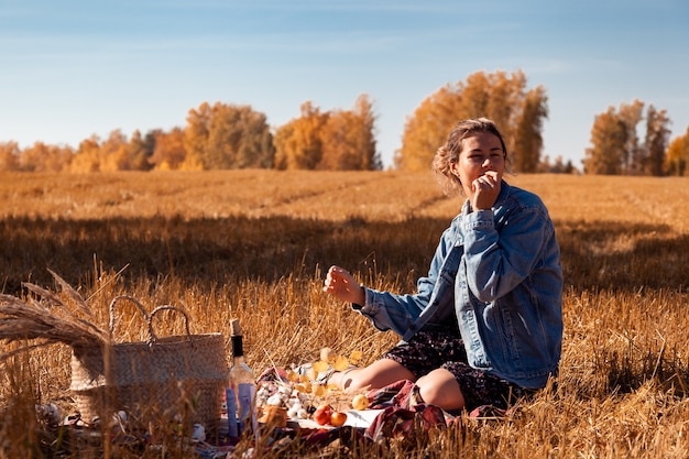 Pique-nique au grand air: une jeune femme vêtue d'une veste en jean et d'une robe mangeant une pomme et profitant de la nature, assise sur un plaid avec un panier de pique-nique, des pommes et du vin.