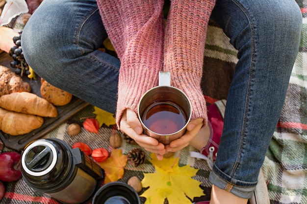 Pique-nique d'automne dans le parc, chaude journée d'automne. La fille tient une tasse de thé dans ses mains. Panier avec des fleurs sur une couverture en feuilles d'automne jaunes. Concept d'automne