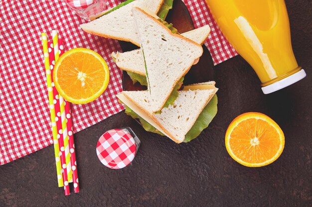 Pique-nique sur une table marron. Nappe à carreaux rouge, corbeille, sandwich aux aliments sains et fruits, jus d'orange. Heure d'été repos. Pose à plat