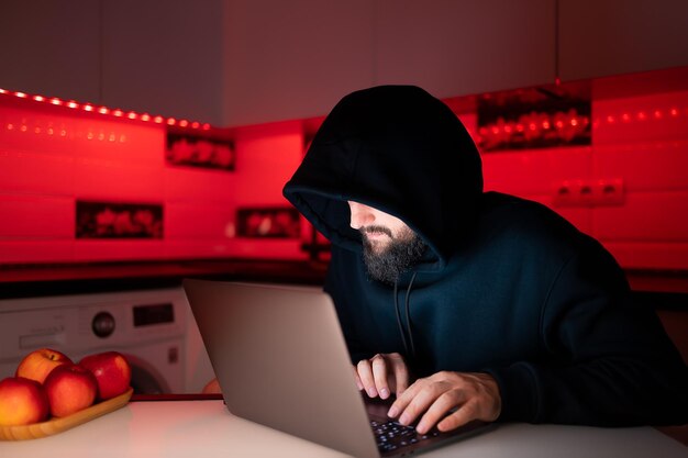 Un pirate dans un sweat à capuche noir avec une capuche sur la tête est assis devant un ordinateur portable et tape sur un clavier